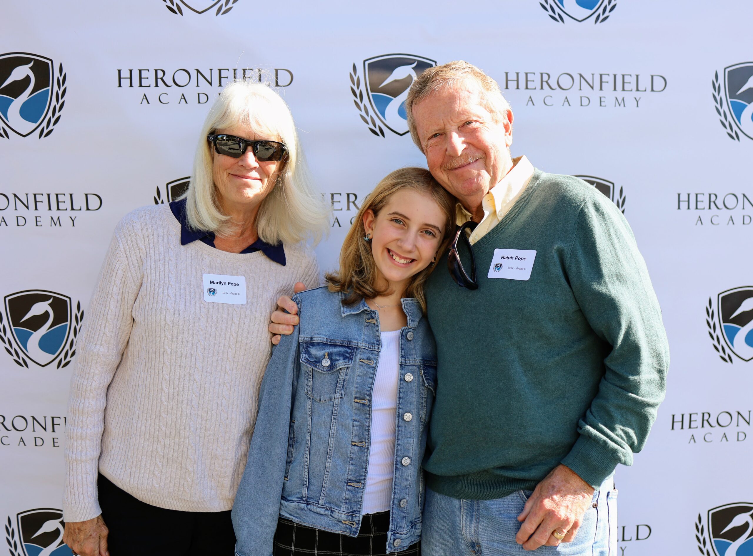 A sixth grader is embraced by her grandparents standing in front of the Heronfield step and repeat banner.
